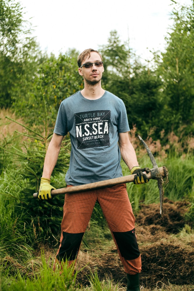 Young man standing in a forest clearing, holding a pickaxe in both hands. The person is wearing sunglasses, a T-shirt, plaid pants and rubber boots.