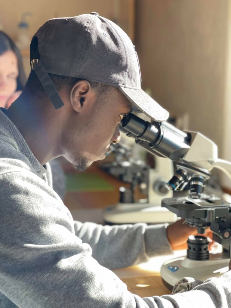 Young man wearing a baseball hat siting at a desk and looking through a microscope.